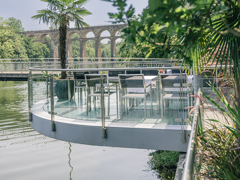 Terrasse am Wasser mit Palmen und historischem Viadukt im Hintergrund.