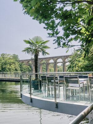 Terrasse am Fluss mit Palme, Fußgängerbrücke und historischem Viadukt im Hintergrund