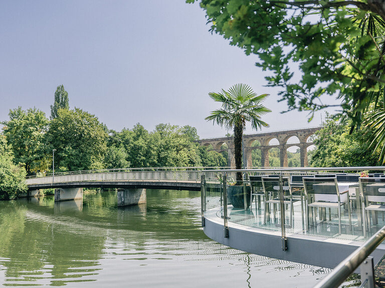 Terrasse am Fluss mit Palme, Fußgängerbrücke und historischem Viadukt im Hintergrund