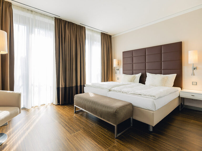 Helles Hotelzimmer mit Doppelbett, großen Fenstern und gemütlicher Sitzecke.